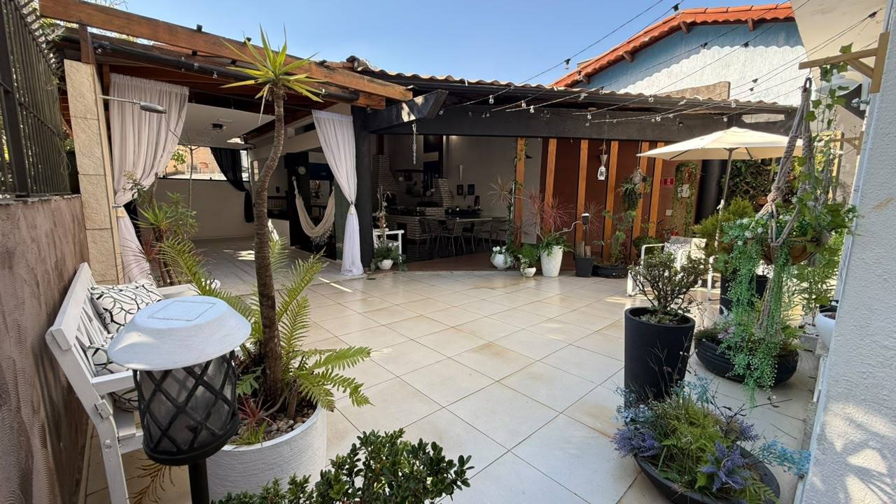 Pátio do Espaço Meneghelli durante o dia, com piso claro, pergolado de madeira, varal de lâmpadas e plantas em vasos